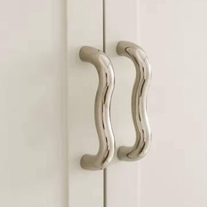 Op de afbeelding: Twee gepolijste, golvende kastgrepen van roestvrij staal. De handgrepen zijn gemonteerd op een witte kastdeur en bieden een modern en strak design. De handgrepen hebben een gebogen, ergonomisch ontwerp.
