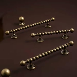 以下が含まれることがあります： アンティーク真鍮キャビネット金具セット。セットには、ビーズのプルハンドルと丸いノブがいくつか含まれています。ハードウェアはヴィンテージの美しさがあり、濃い茶色の表面に表示されています。ハンドルは約15cmです。