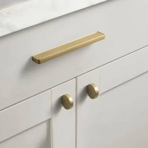 Puede incluir: Primer plano de un armario blanco con herrajes dorados. El armario presenta un tirador largo y rectangular y dos pomos redondos. Los herrajes tienen un acabado cepillado, lo que añade un toque de elegancia al diseño.