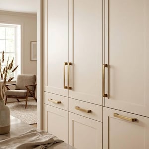 May include: Cream kitchen cabinets with gold-coloured handles. The cabinets have a modern design with rectangular panels and long, vertical handles. A marble countertop and a vase with dried plants are in the foreground.