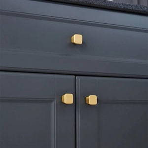 Könnte beinhalten: Drei goldene quadratische Schrankknöpfe an einem dunkelgrauen Schrank. Die Knöpfe sind etwa 2,5 cm quadratisch.