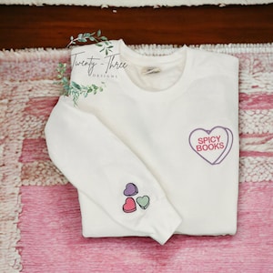 Puede incluir: Sudadera blanca con un gr&aacute;fico de coraz&oacute;n morado que dice "Spicy Books" y tres corazones m&aacute;s peque&ntilde;os en la manga. La sudadera est&aacute; doblada sobre una alfombra rosa y blanca.