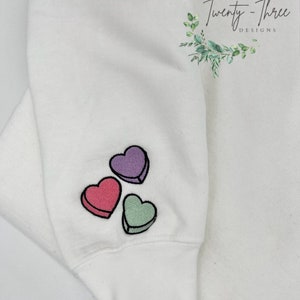 Puede incluir: Sudadera blanca con tres corazones bordados en rosa, morado y verde. Los corazones tienen la forma de corazones de conversaci&oacute;n de caramelo. La sudadera tiene un pu&ntilde;o en la parte inferior de la manga.