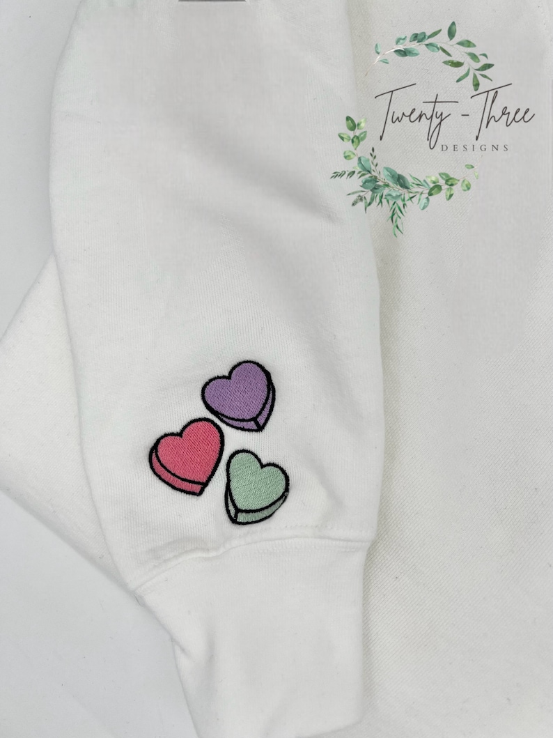 Puede incluir: Sudadera blanca con tres corazones bordados en rosa, morado y verde. Los corazones tienen la forma de corazones de conversaci&oacute;n de caramelo. La sudadera tiene un pu&ntilde;o en la parte inferior de la manga.