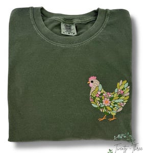 May include: An olive green Comfort Colors t-shirt with an embroidered floral chicken design on the front. The chicken features pink, yellow, and green floral details. The shirt has a crew neck and the size is L-G.