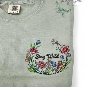 May include: A light green t-shirt with a floral design and the text "Stay Wild" in black.