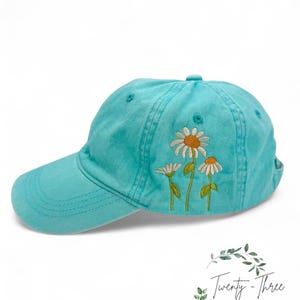 May include: A turquoise blue baseball cap with embroidered white daisies. The daisies have yellow centers.