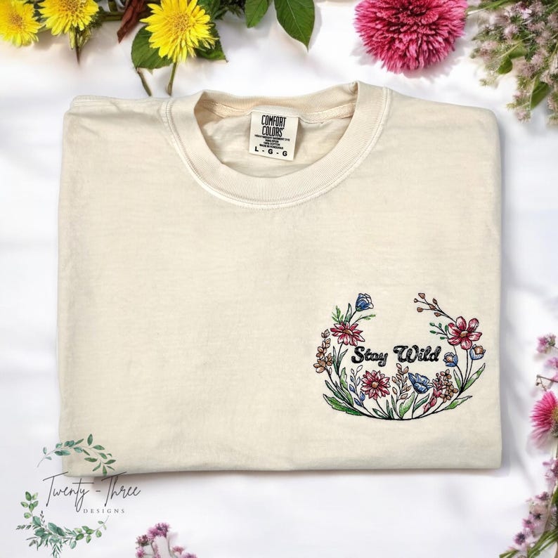 May include: A cream colored t-shirt with a floral embroidery design that says "Stay Wild".