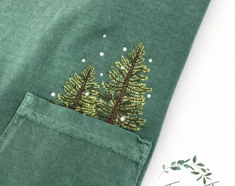 Könnte beinhalten: Ein grünes T-Shirt mit einer Tasche, die mit einem gestickten Design aus drei Kiefern mit weißen Schneeflocken verziert ist. Der Text "Twenty-Three Designs" ist auf einem Kranz aus grünen Blättern in der unteren rechten Ecke gestickt.
