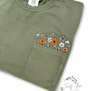 Puede incluir: Una camiseta verde oliva con un bolsillo en el pecho bordado con un grupo de flores naranjas, blancas y marrones con tallos y hojas verdes. La camiseta tiene una etiqueta Comfort Colors en el cuello y el logo Twenty-Three Designs.