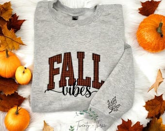 Puede incluir: Una sudadera gris con las palabras "FALL vibes" en un diseño de cuadros marrones. Una pequeña hoja está bordada en la manga. Las calabazas y las hojas de otoño rodean la sudadera.