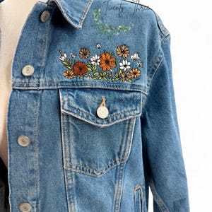May include: Light blue denim jacket with embroidered floral design in orange, white, and brown. The jacket has a classic button-up front, chest pockets, and the text "Twenty-Three" on the upper right shoulder.