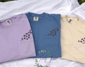 May include: Three t-shirts with a pocket on the front. Each shirt has a different color: lavender, blue, and beige. The pocket on each shirt has a design of butterflies.