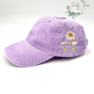 Op de afbeelding: Een lavendelkleurige baseballpet met geborduurde witte madeliefjes. De pet heeft een licht verweerde look en is gemaakt van een zachte, comfortabele stof.