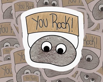 Puede incluir: Una roca gris con ojos saltones que lleva un cartel de madera que dice "YOU Rock!" El cartel está sujeto a la roca con una cuerda blanca.