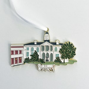 Puede incluir: Adorno de metal dorado que representa un edificio blanco con una torre de reloj y árboles verdes. El edificio tiene un techo rojo y el texto "Oxford" está escrito debajo del edificio.