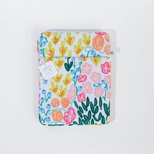 Könnte beinhalten: Eine weiße Canvas-Tech-Hülle mit einem bunten Blumenmuster. Die Hülle hat einen Reißverschluss und einen Riemen. Der Text "Anna Whitlam Co." ist auf einem an der Hülle befestigten Etikett aufgedruckt.