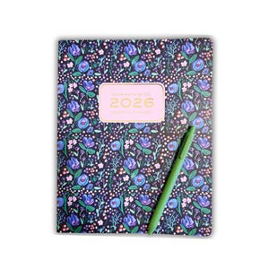Könnte beinhalten: Ein monatlicher Planer für 2026 mit floralem Muster, dunklem Hintergrund und einem rosa Etikett mit der Aufschrift "ANNA WHITMAN CO. 2026 MONTHLY PLANNER". Ein grüner Stift liegt auf dem Planer. Das Blumenmuster umfasst blaue, violette und weiße Blumen.
