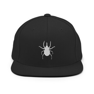 May include: Black flat-brimmed baseball cap with a white embroidered beetle design on the front. The cap has a structured fit and a curved brim. The beetle design is detailed and centered on the cap.