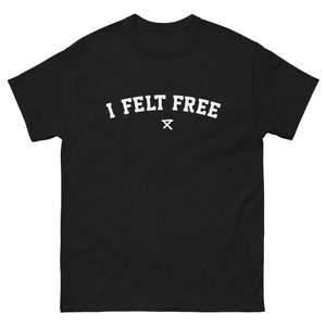 Puede incluir: Camiseta negra con el texto blanco "I FELT FREE" en forma de arco. Debajo del texto hay un pequeño símbolo blanco. La camiseta está hecha de una tela gruesa y duradera.