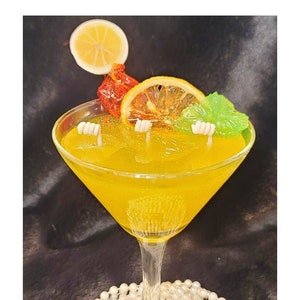Puede incluir: Una vela de vidrio de martini amarillo con tres mechas, decorada con rodajas de limón, un caramelo rojo y una hoja verde. La vela está sentada sobre un collar de perlas blancas.