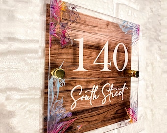 May include: A square acrylic house number sign with a woodgrain background and colorful floral accents. The number 140 is printed in white with the text "South Street" below it.