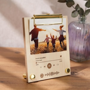 Può includere: Una cornice per foto in legno con una clip dorata e una foto di una famiglia di quattro persone che corrono in un campo. La cornice ha un design di lettore musicale con il testo "You're beautiful" e "James Blunt" su di essa.