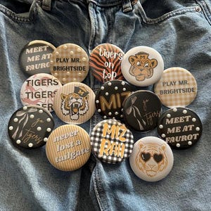May include: A collection of round buttons with various designs and text, including "Tigers on Top," "MIZ RAH," and tiger-themed graphics. The buttons are arranged on a denim background, showcasing a variety of colors and patterns.