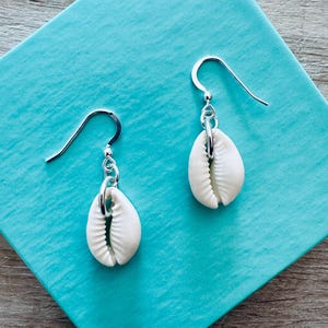May include: A pair of silver earrings with white cowrie shell charms. The earrings have a simple hook design.