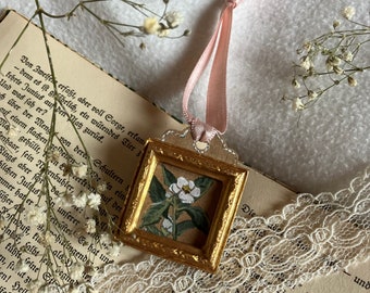 May include: A gold-framed picture ornament with a white flower and green leaves painted inside. The ornament is hanging from a pink ribbon and is resting on a book with white lace.