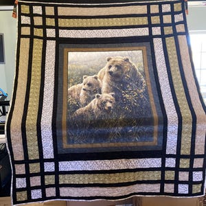 Puede incluir: Un edredón grande con una imagen central de tres osos pardos en un entorno forestal. El edredón tiene un borde con un patrón de cuadrícula en negro, beige y blanco. El diseño es un artículo de decoración del hogar con temática de la naturaleza.