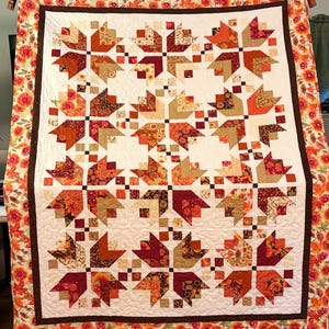 Op de afbeelding: Een handgemaakte quilt met een herhalend patroon van gestileerde esdoornbladeren in tinten oranje, bruin en beige. De bladeren zijn geplaatst tegen een witte achtergrond en omlijst door een bloemenrand. De quilt is een gezellig, herfstthema woondecoratie-item.
