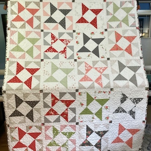 Op de afbeelding: Een quilt met een patchwork-ontwerp met een herhalend patroon van rode, groene en grijze sterren op een witte achtergrond.