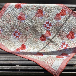 Puede incluir: Una colcha blanca y roja con un patrón de corazón. La colcha está doblada y sentada en un banco de madera.