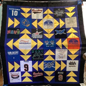 May include: A handmade quilt with a blue and gold starburst pattern. The quilt features squares with baseball-themed logos, team names, and the numbers 10 and 9. The quilt is bordered with black fabric.