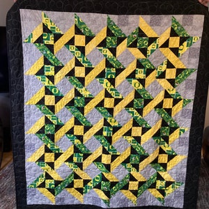 Pode incluir: Um edredom preto, amarelo e verde com um padrão geométrico. O edredom é feito de tecido com um padrão repetido da letra "O" e um design verde e amarelo.