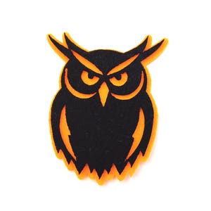 May include: A black and orange felt owl cutout. The owl has a black body with orange outlining the eyes, beak, and edges. The owl has pointed ear tufts and a stern expression. The cutout is likely for a Halloween or fall craft project.