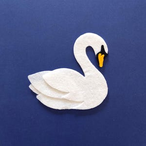 Könnte beinhalten: Ein weißer Filzschwan mit einem schwarzen und gelben Schnabel. Der Schwan befindet sich auf einem blauen Hintergrund.