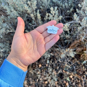 Könnte beinhalten: Ein Miniatur-Auflaufform in Weiß mit Deckel und blauem Blumendesign. Das kleine Gericht wird in einer Hand gehalten, mit einem blauen Ärmel in der Ecke. Der Hintergrund ist verschwommen und zeigt Blätter.