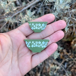 May include: Two miniature green bowls with white floral patterns. The bowls are small, likely for decorative purposes or dollhouses. The design is reminiscent of vintage kitchenware.