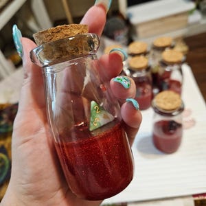 May include: A clear glass bottle with a cork stopper, filled with red glitter and a small, green and white die. Several similar bottles are in the background. The bottle is held in a hand with long, painted nails.
