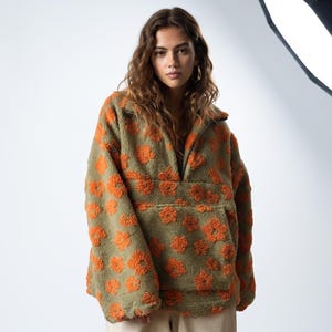 May include: A long-sleeved, olive green fleece pullover featuring a textured orange floral pattern throughout. The garment includes a high collar with a partial front zipper, dropped shoulders, and a large, horizontal front kangaroo pocket.