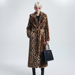 May include: A long, belted faux fur coat featuring a brown and black leopard print pattern, paired with a structured black leather handbag with gold-tone hardware, and black leather boots. The coat has a wide collar and long sleeves.