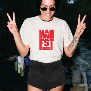 May include: A cream-coloured short-sleeve t-shirt featuring the word MANIFEST printed in bold red capital letters on the chest. The design is arranged in a block-like layout with the letters stacked vertically and horizontally in a modern font.