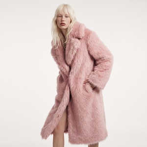 May include: A long, light pink faux fur coat featuring a wide notched lapel collar, long sleeves, and a straight-cut silhouette that falls to the mid-thigh. The garment is constructed with a thick, textured pile and includes side pockets.