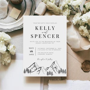 May include: A wedding invitation with the names "Kelly and Spencer" in elegant black font. The invitation includes the date, time, and location of the ceremony, along with a mountain and tree illustration at the bottom. The invitation is on a white background.