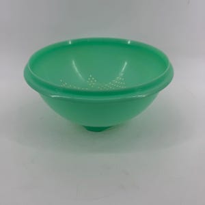May include: A mint green plastic colander bowl with a wide, rounded rim and a series of small, circular drainage holes arranged in a pattern at the base. The bowl features a smooth, semi-translucent finish and a small, centered base support.