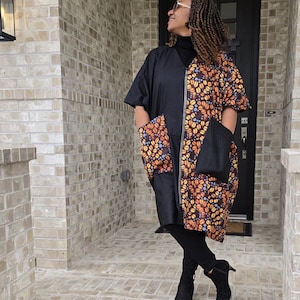 May include: A black and orange patterned dress with a zipper down the front. The dress has two large pockets on the sides. The person wearing the dress is standing on a brick patio.