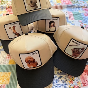 May include: A collection of beige baseball caps with black brims. Each cap features a square patch with a dog portrait and a name: Nash, Truman, Navy, Otis, and Susan. The caps are arranged on a colorful patchwork quilt.
