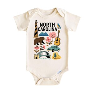 May include: Cream-colored baby bodysuit with a colorful graphic of North Carolina landmarks, including a lighthouse, mountains, bear, flowers, guitar, car, bridge, and a bird. The text "NORTH CAROLINA" is printed in black. Made of soft material.